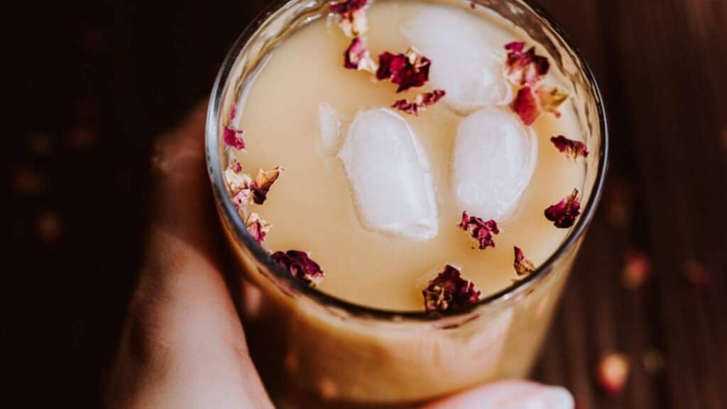 A hand holds a clear glass filled with an iced beverage scattered with rose petals.