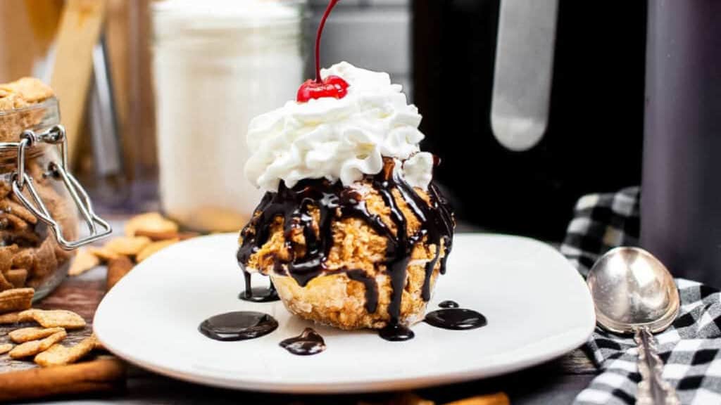 Low angle shot of air fryer fried ice cream on a white plate topped with whipped cream, chocolate sauce, and a maraschino cherry.