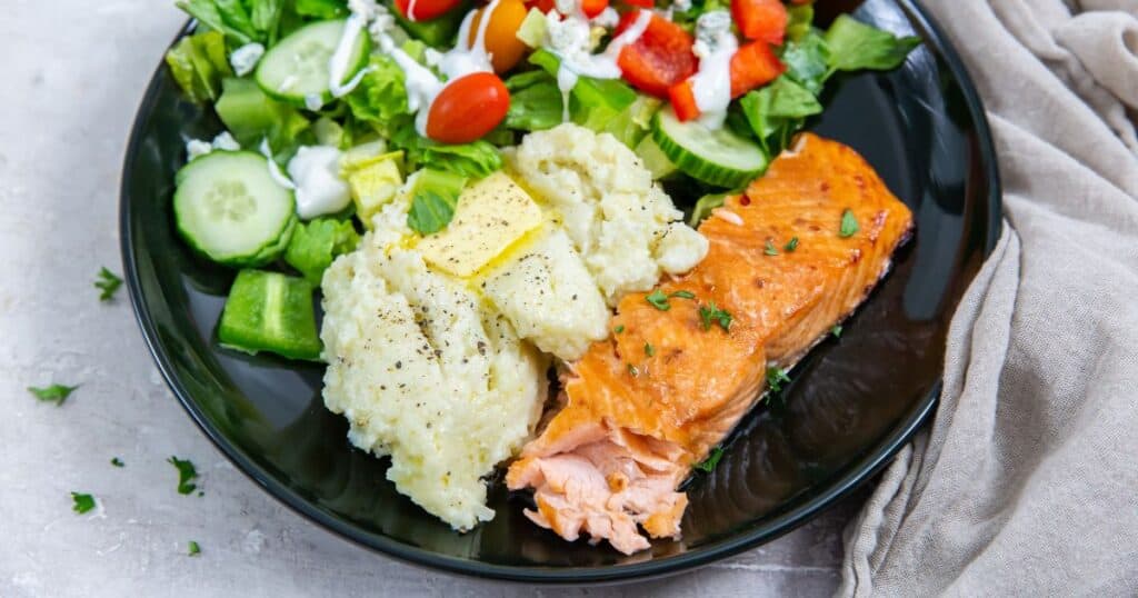A balanced plate with air-fried salmon, mashed potatoes, and a fresh salad with greens, cucumbers, tomatoes, and feta cheese, served on a black plate.