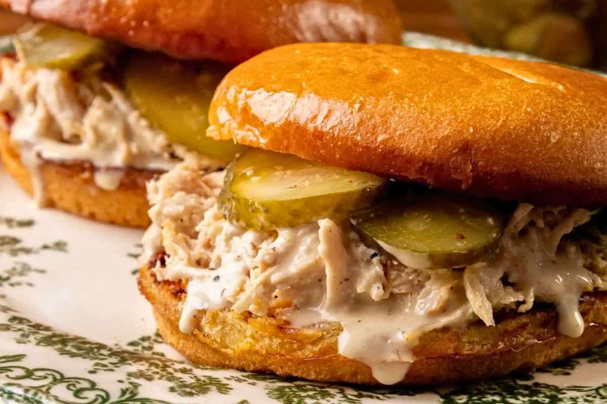 Two Alabama Chopped Chicken Sandwiches with pulled chicken, creamy Alabama white BBQ sauce, and pickle slices on shiny, toasted buns, served on a decorative plate.