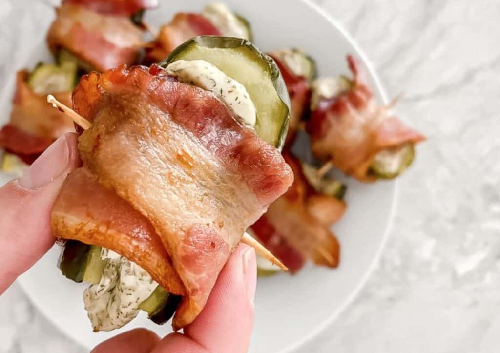 A hand holding a bacon-wrapped pickle bite skewered with a toothpick.