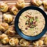Bacon beer cheese in a dark bowl surrounded by pretzel knots on a cutting board.