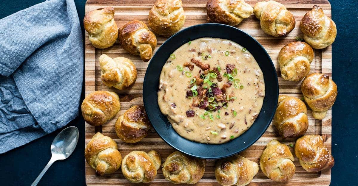 Bacon beer cheese in a dark bowl surrounded by pretzel knots on a cutting board.