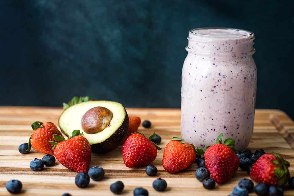A berry avocado smoothie in a glass mason jar with strawberries, blueberries and avocado on the side.