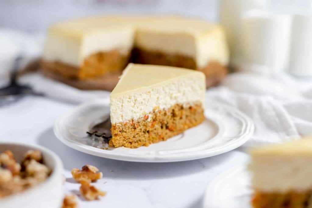 Slice of double decker carrot cake on a plate with the rest of the cake in the blurred background.