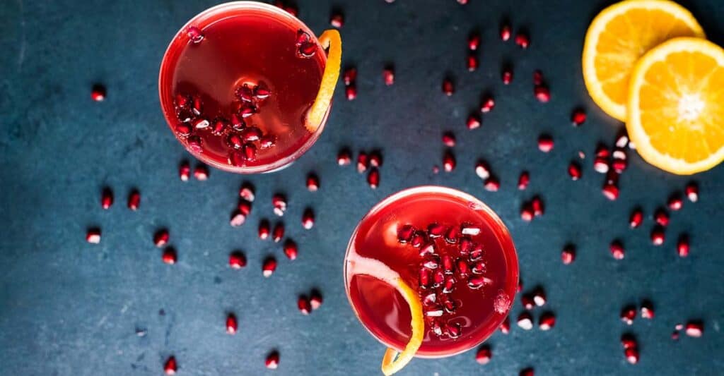 Overhead shot of two champagne pomegranate cocktails in glass goblets with pomegranate arils and orange zest.