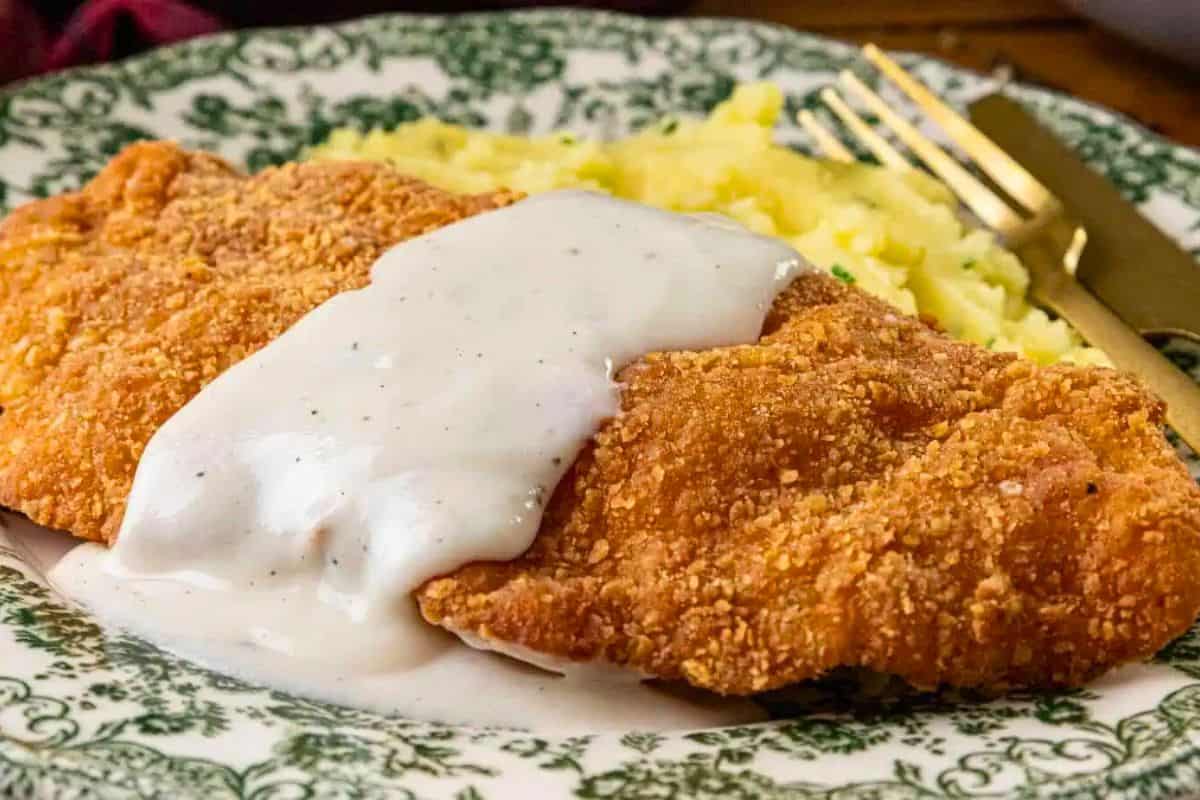 A classic Southern comfort food, this Chicken Fried Chicken features a breaded and fried cutlet topped with rich gravy, served with mashed potatoes on a decorative green and white plate beside a gold fork and knife.