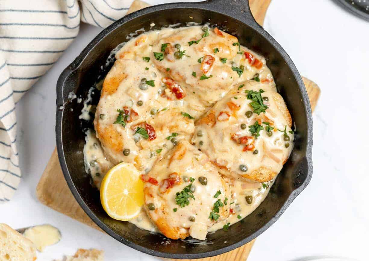 A cast iron frying pan with chicken piccata and a lemon wedge on the side on a white background.