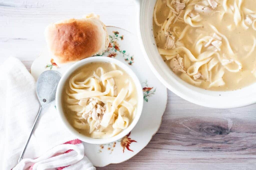 A bowl of chicken and noodles with a roll next to it and the pot of soup next to it.