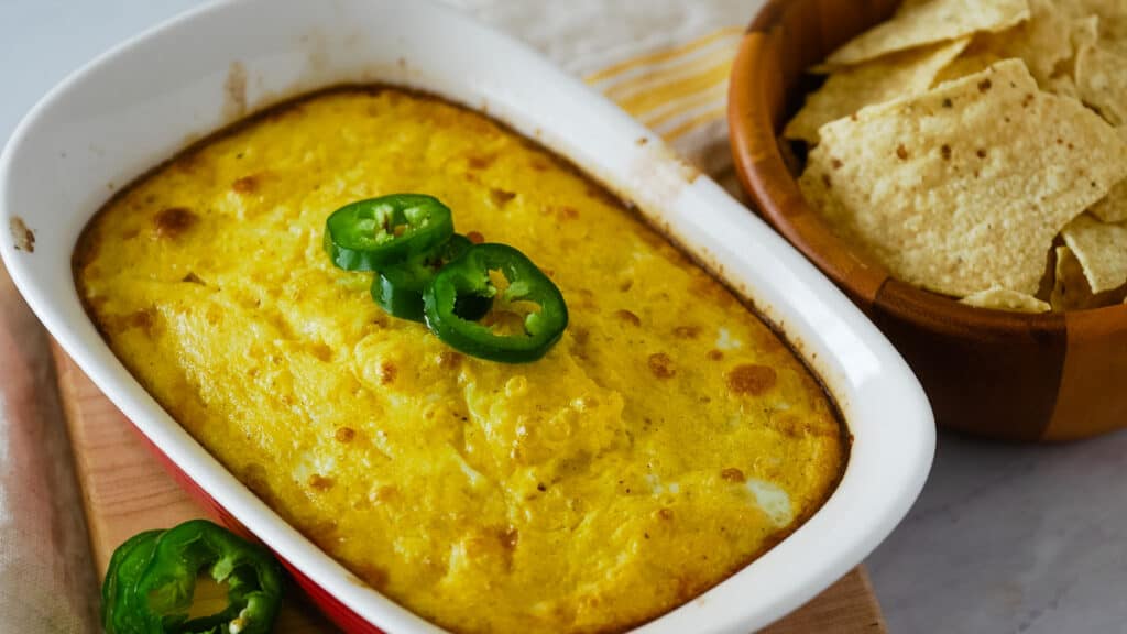 Chile relleno casserole topped with jalapenos in a red dish next to a bowl of chips.