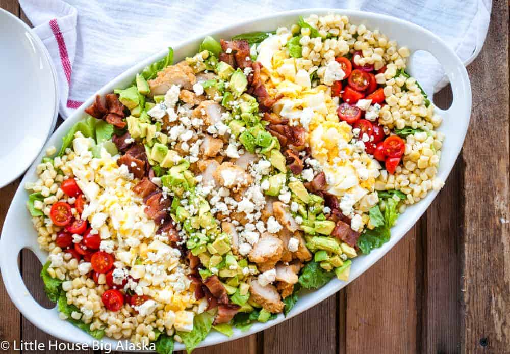 A big cobb salad on a platter with crispy chicken, avocados, eggs, tomatoes, all on top of greens.