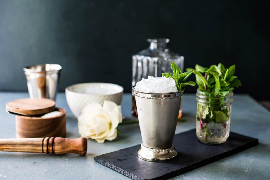 A mint julep in a silver mint julep cup with sprigs of mint and a decanter of bourbon and other ingredients in the background. 