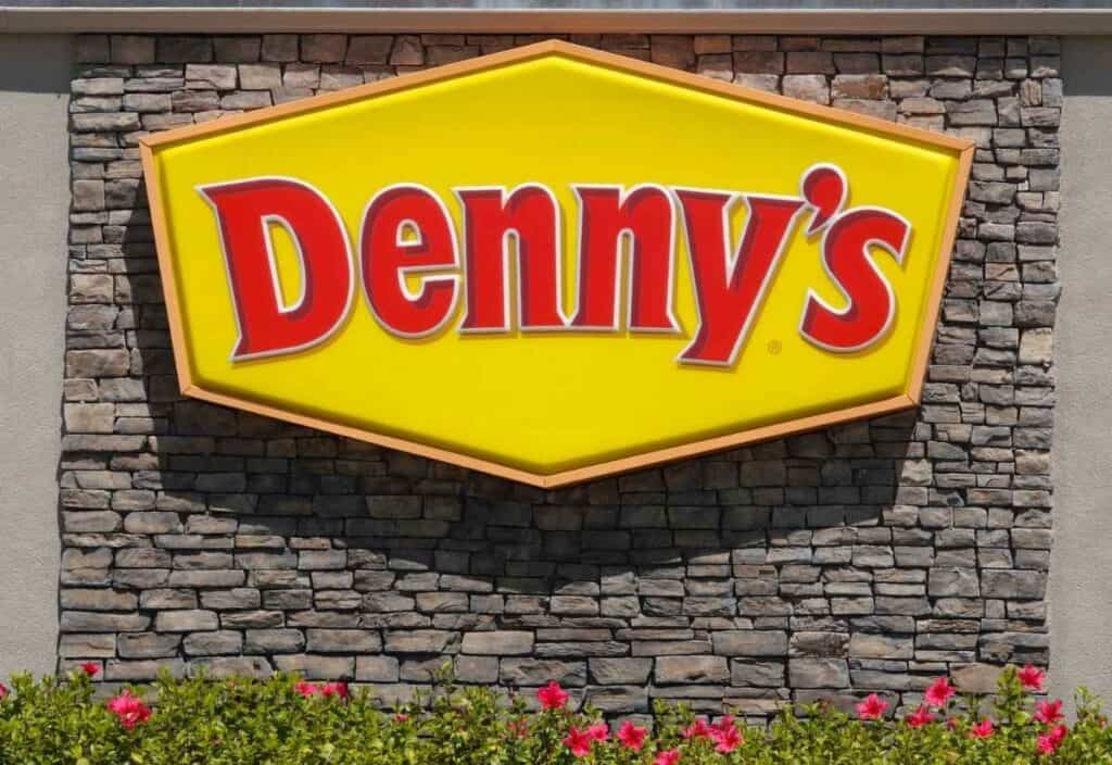 A yellow and red Denny's restaurant sign is mounted on a stone wall above green bushes with pink flowers.