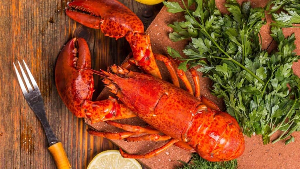 Cooked whole lobster on a wooden board with fresh parsley, dill, lemon slices, and a fork on the side&mdash;perfect for those curious about how to cook lobster and present it beautifully.