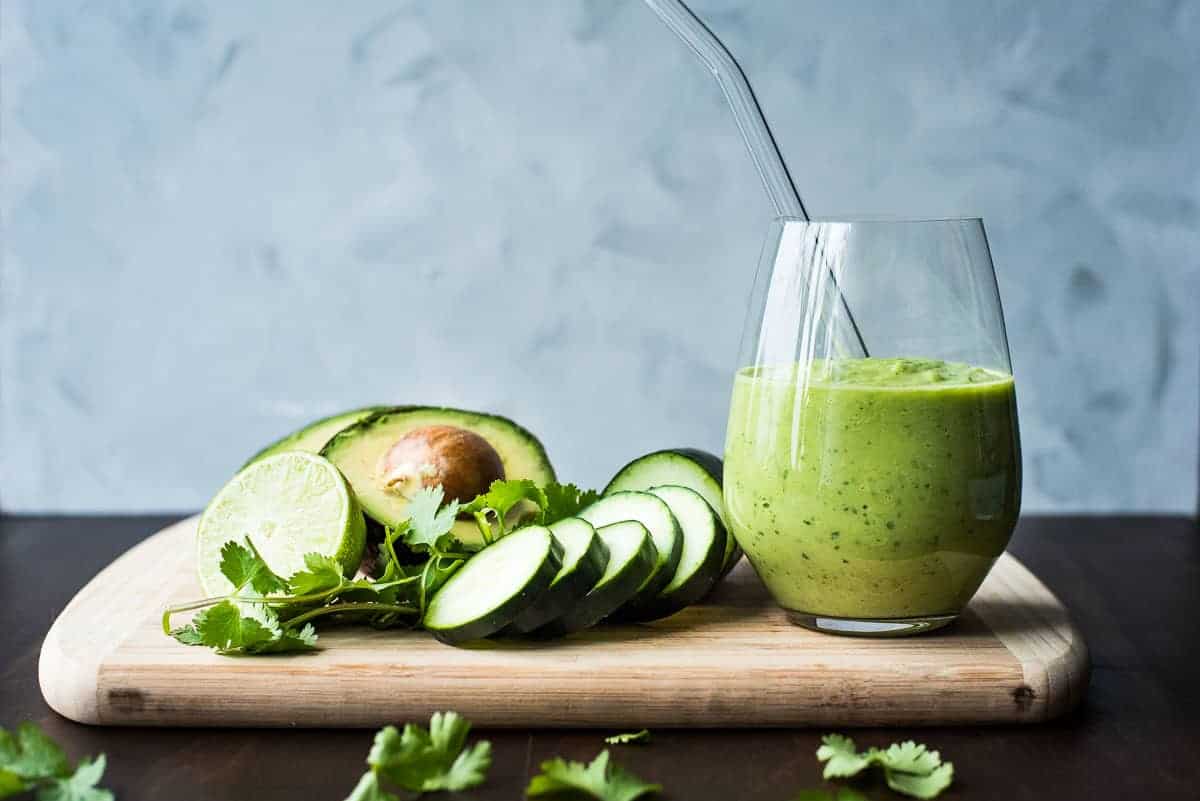 A green smoothie with cucumbers, cilantro and avocado on a wooden cutting board.