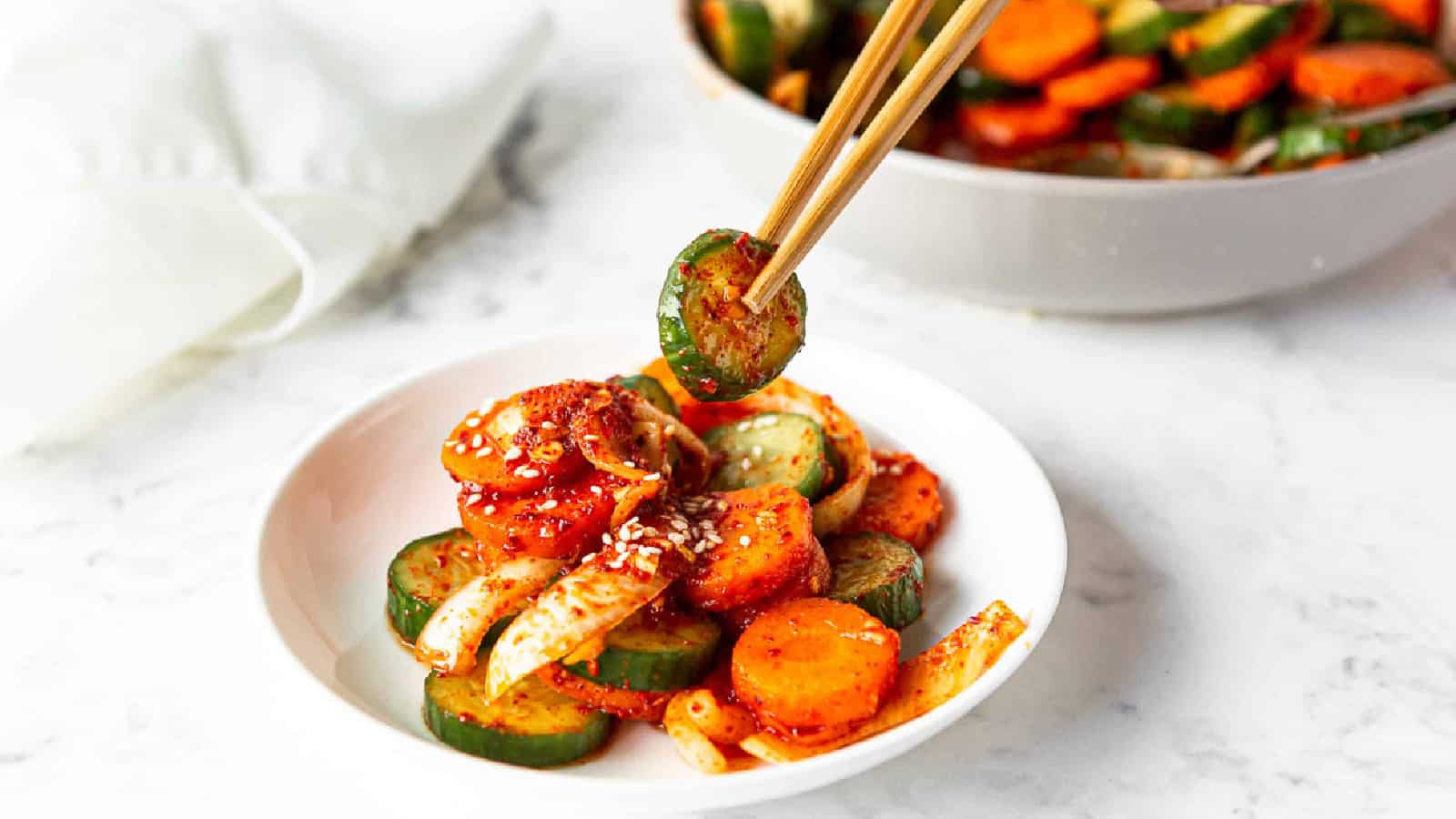 Cucumber kimchi on a white plate with a pair of chopsticks lifting a slice of cucumber.