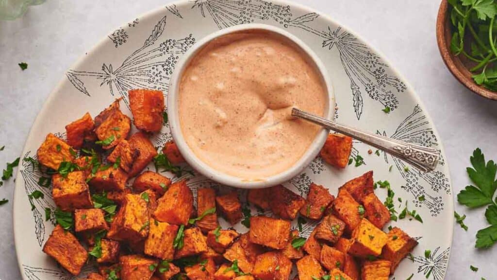 Dipping sauce in a bowl with a spoon in it and sweet potato cubes surrounding it. 