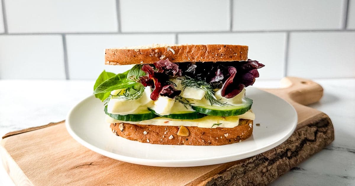 An egg and cucumber sandwich with pepper jack cheese, spring mix, and dill sauce is shown on a white plate over a rustic wooden cutting board.