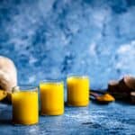 Ginger Turmeric Shots on a blue board with jug behind.