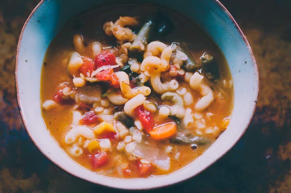 A small ceramic bowl filled with pasta and vegetables suspended in a red broth.