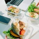 Three containers of assorted healthy food on a desk with a laptop, smartphone, and potted plant; a hand reaches for salad in one container.