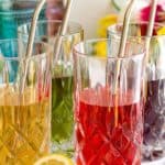 Four tall glasses with metal straws hold vibrant drinks—iced tea in yellow, red, and purple hues—set on a table adorned with fruit and flowers in the background.