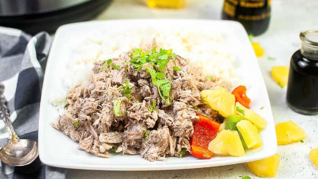 Kalua pork on a white plate with rice , pineapple, and red pepper.