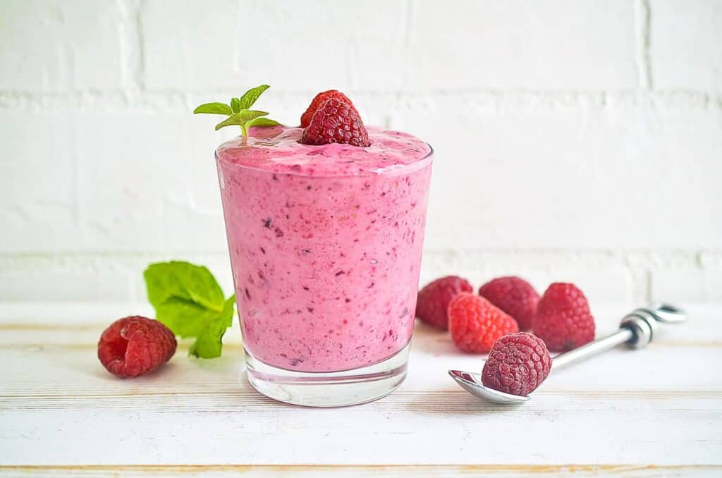 A raspberry smoothie in a glass with a spoon and raspberries nearby.