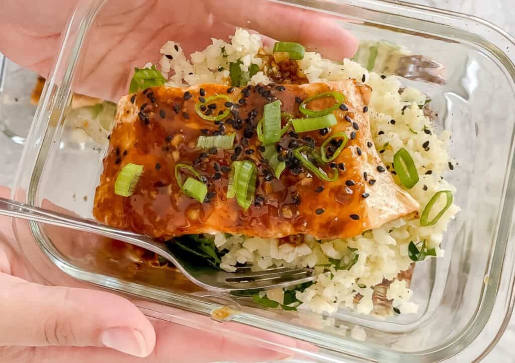 Meal prep saucy salmon with parsley cauliflower rice in a glass container being held in hands.