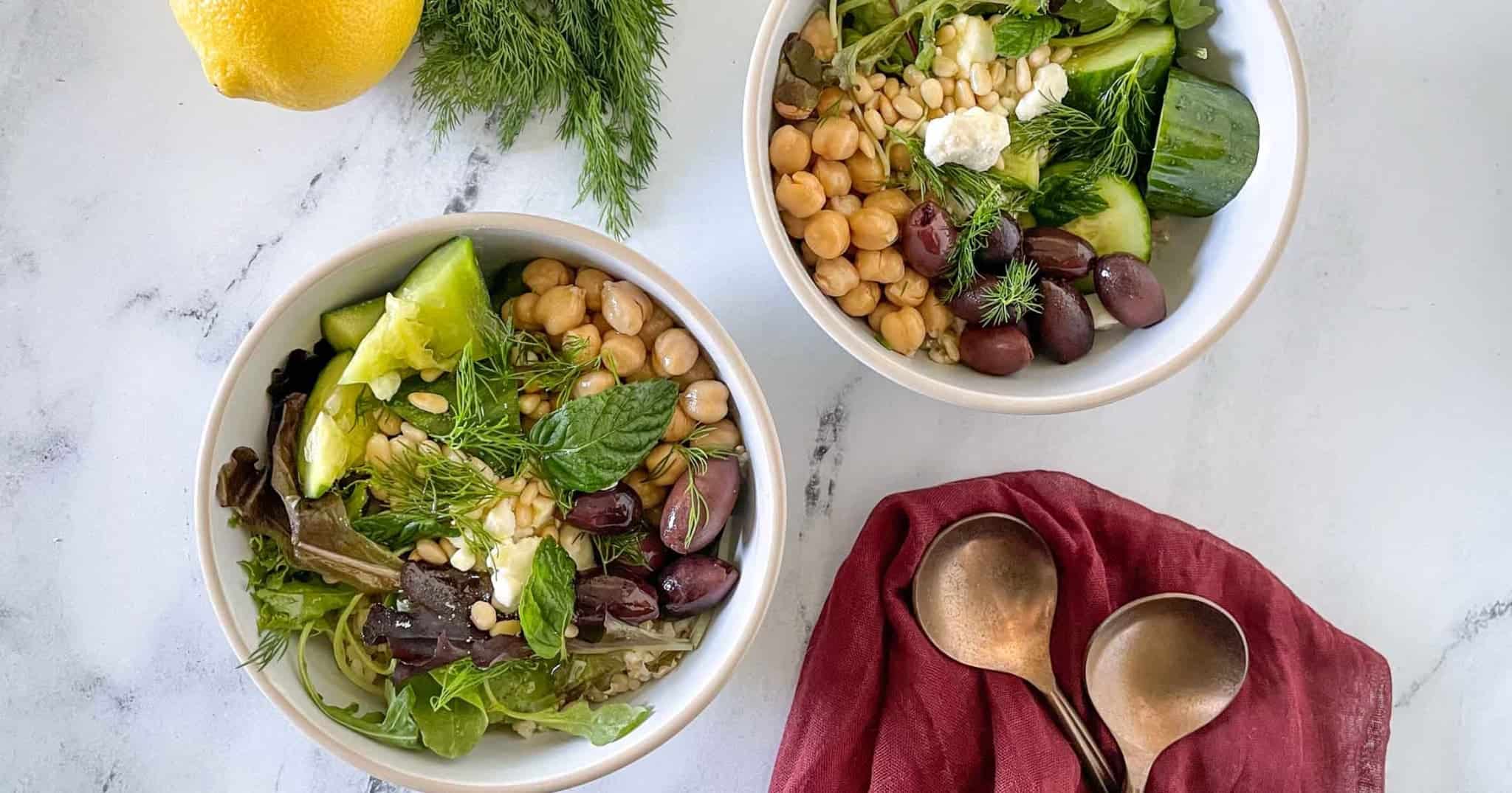 Two grain bowls are shown on a white marble counter with forks, a red linen, dill, and a lemon.