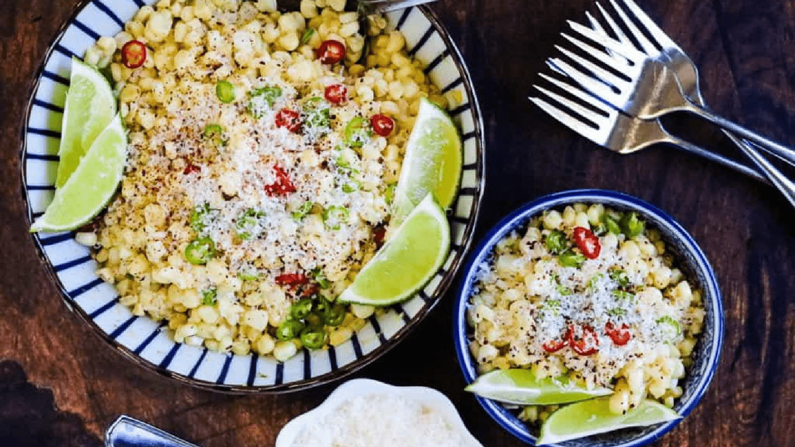 Mexican corn salad in bowls, garnished with parmesan cheese and lime wedges, with forks on the side.
