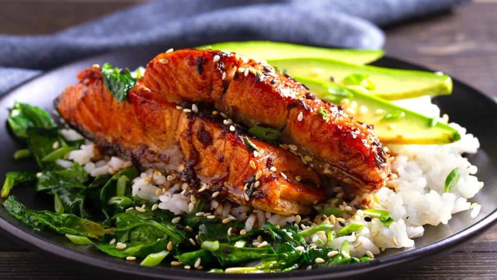 Miso salmon on a black plate with rice, sliced avocado, spinach and sesame seeds.