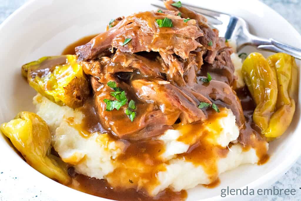 Mississippi Pot Roast over mashed potatoes with gravy in a white bowl.