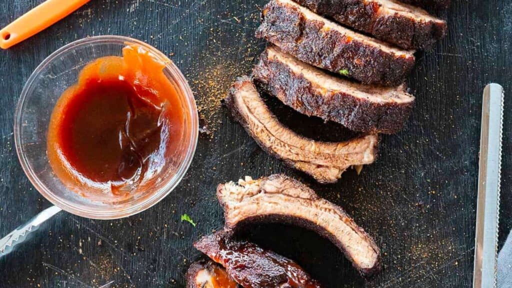 sliced baby back ribs by bowl of BBQ sauce.