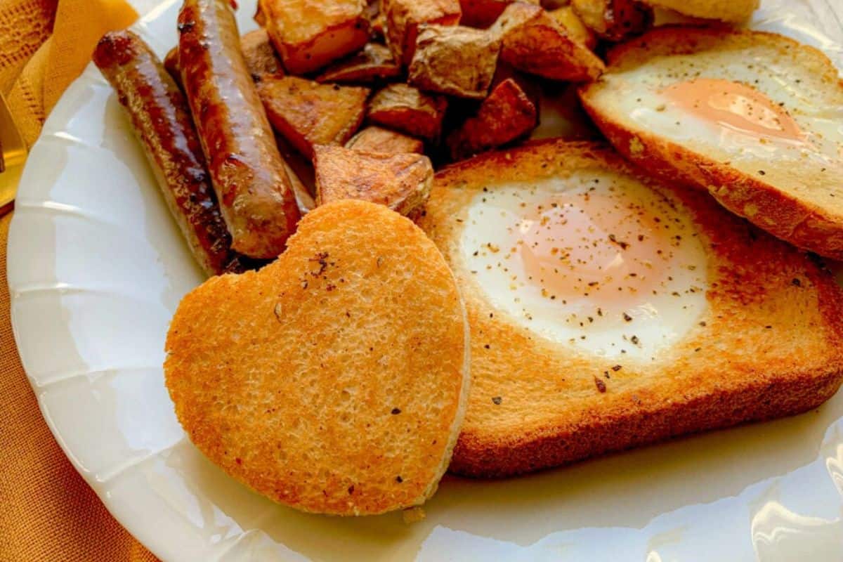 A plated breakfast featuring heart-shaped toast with a sunny-side-up egg in the center, seasoned with pepper. Accompanied by sausages, roasted potatoes, and another toast with an egg, all on a white plate with a yellow napkin nearby.