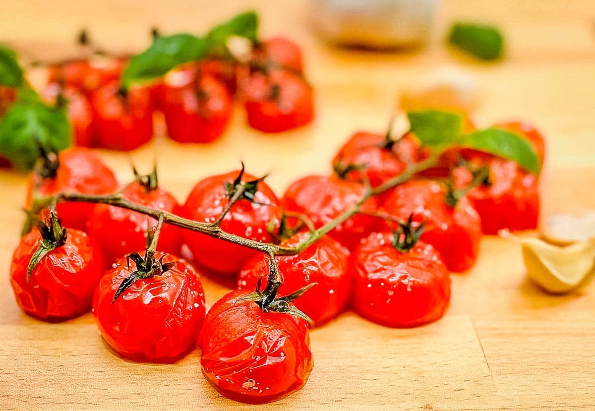 A closeup shot of roasted cherry tomatoes on the vine on a wooden cutting board surrounded by basil and garlic.