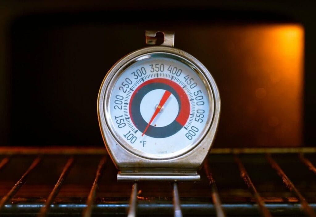 A metal oven thermometer on an oven rack shows a temperature reading of about 375°F, with the oven interior illuminated in the background.