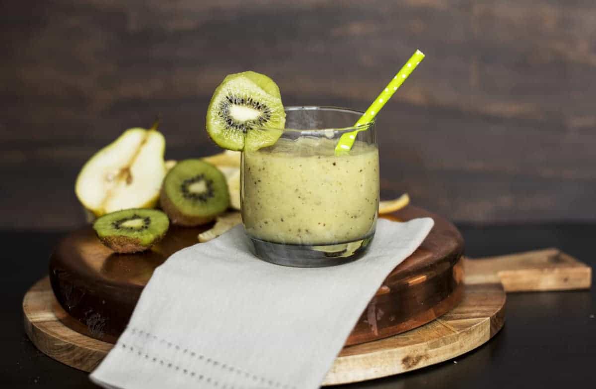 A kiwi pear green smoothie in a glass with a kiwi on the rim and a green straw.