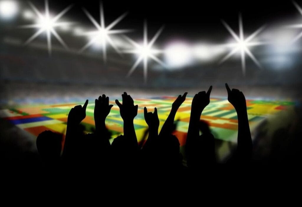 Silhouettes of cheering fans with raised hands in a stadium, bright lights shining overhead, international flags visible on the field in the background.