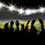 Silhouettes of cheering fans with raised hands in a stadium, bright lights shining overhead, international flags visible on the field in the background.