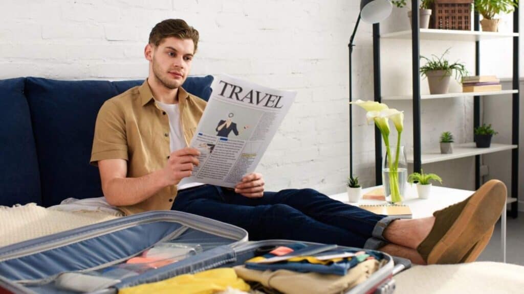 A man sits on a bed reading a travel newspaper, with an open suitcase and neatly folded clothes in front of him in a bright, modern room.