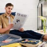 A man sits on a bed reading a travel newspaper, with an open suitcase and neatly folded clothes in front of him in a bright, modern room.