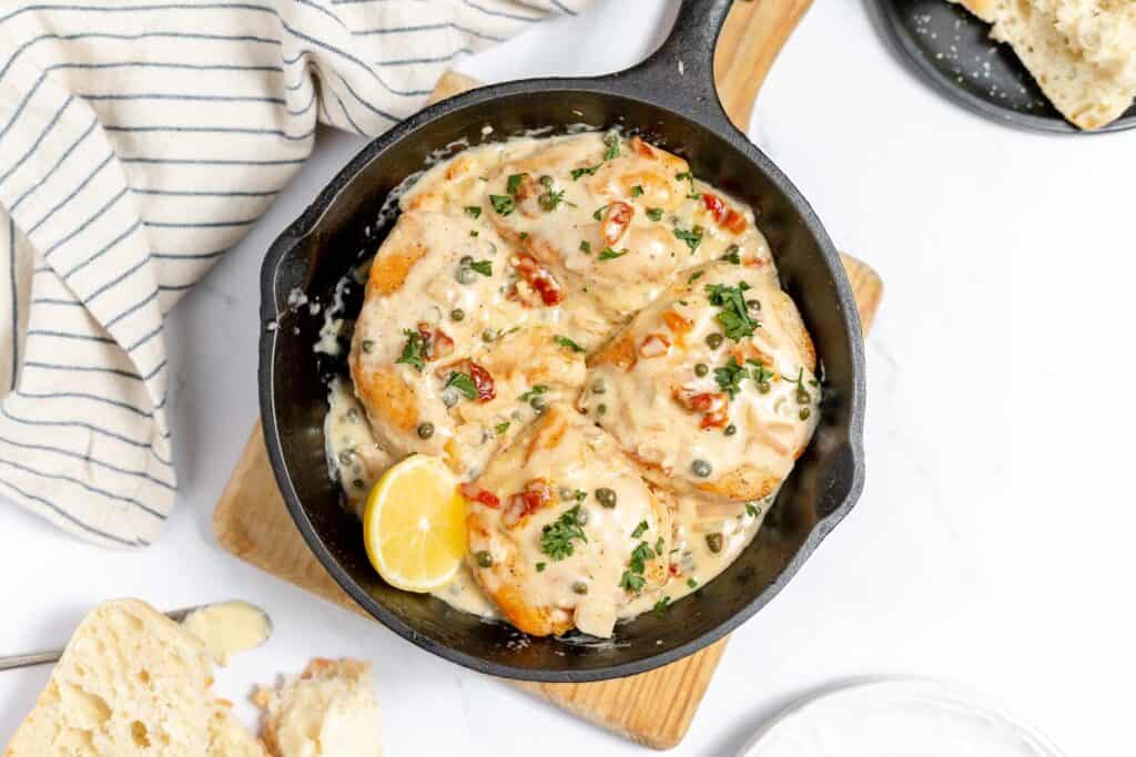 A skillet with chicken piccata in it on a wooden board on a white back ground.