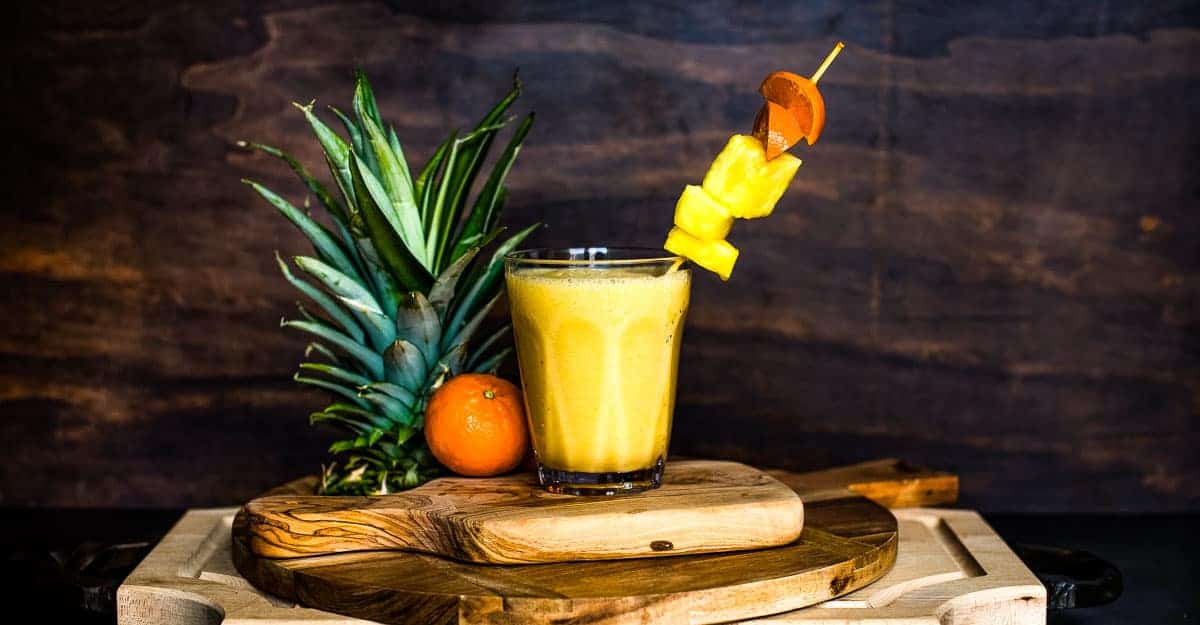 A smoothie in a glass on a stack of cutting boards next to a fresh orange and pineapple top with text overlay.