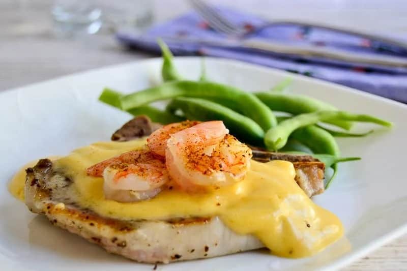 Bone in pork chop topped with hollandaise sauce and shrimp, on a white plate next to green beans.
