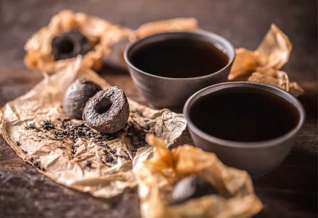 Pu-erh tea in its dried form as well as brewed form in dark ceramic tea cups.