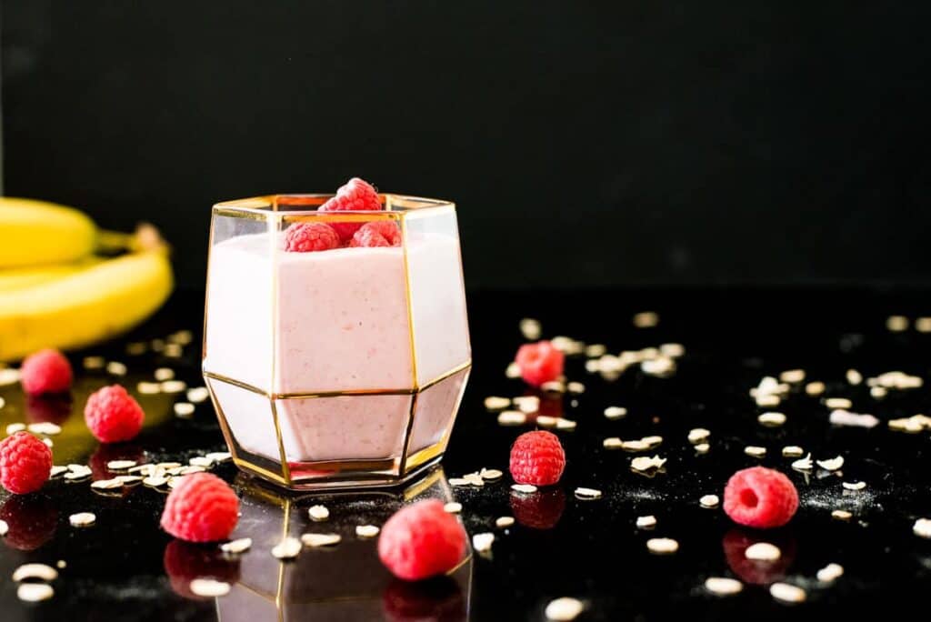 Raspberry smoothie with raspberries on top in gold-trimmed glass on a black surface.