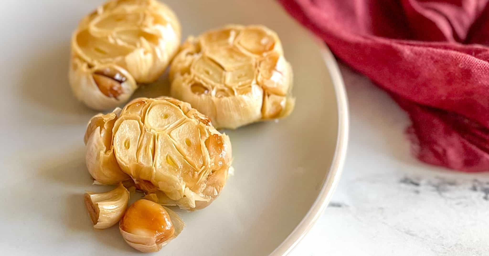 Three heads of roasted garlic sit on a white plate with a red linen in the background.