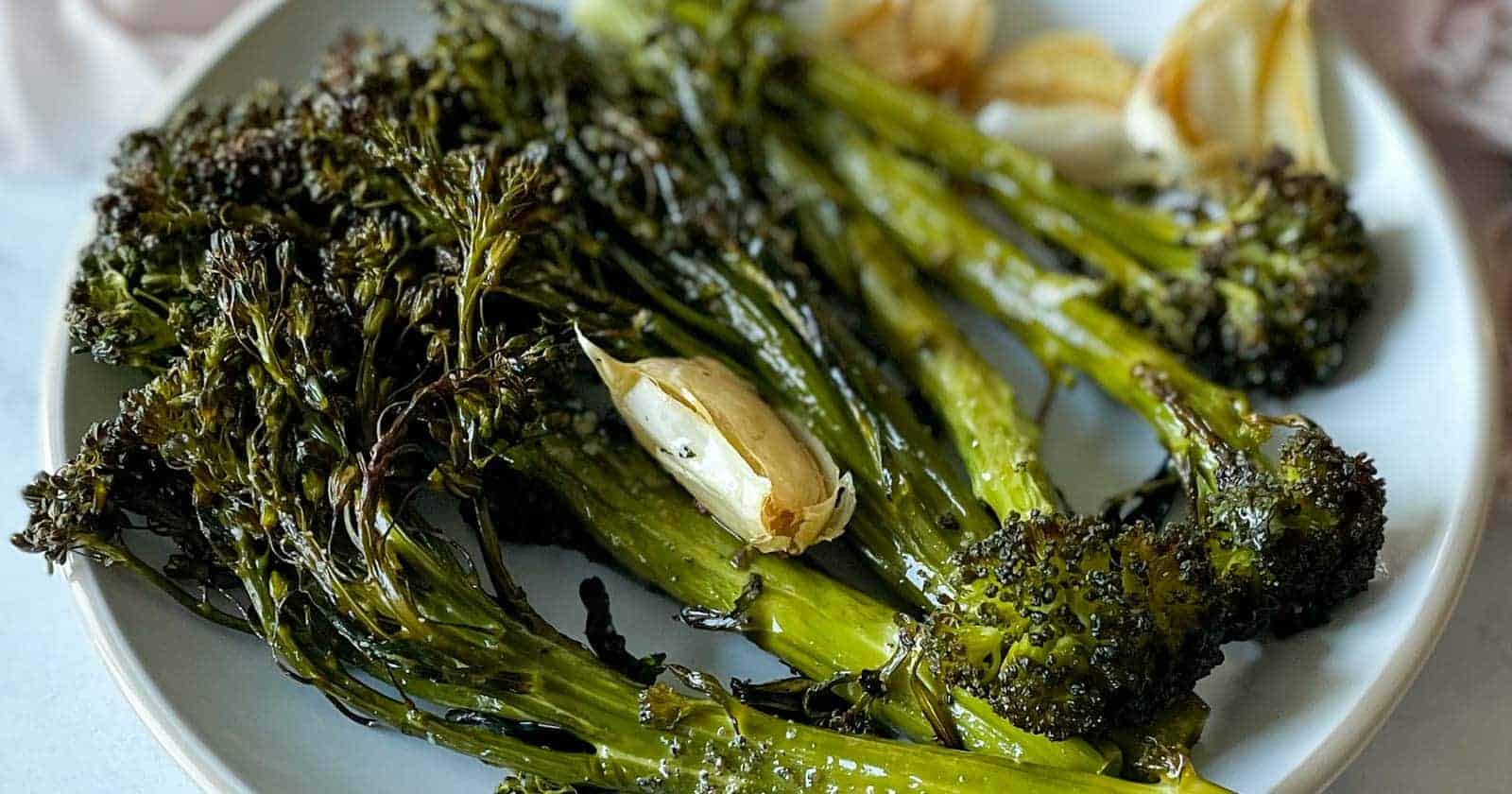 A closeup of roasted tenderstem broccoli and garlic are shown on a white plate.