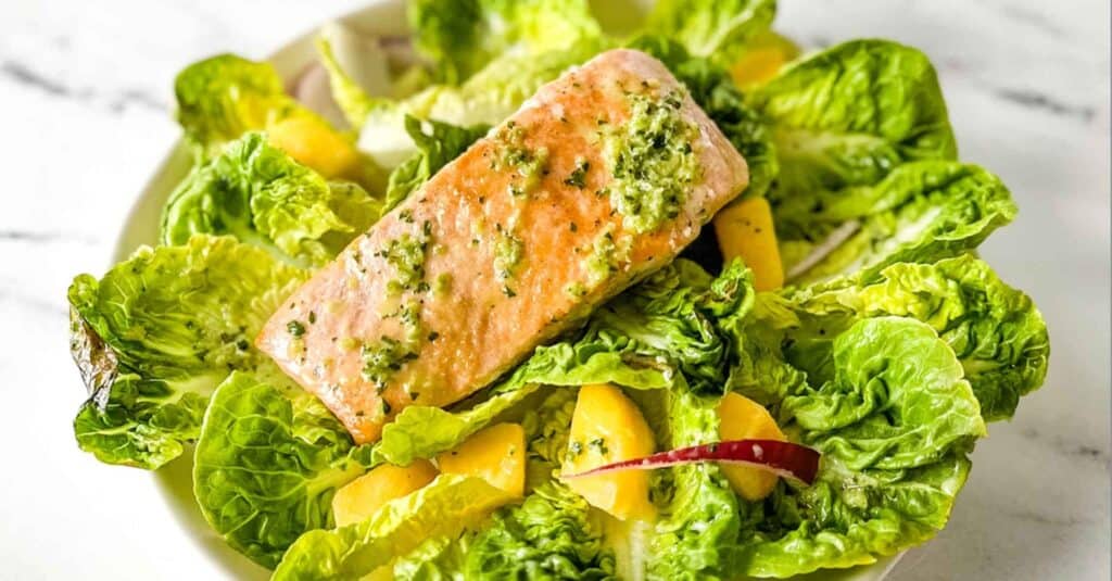 A fillet of salmon sits on a bed of little gem lettuce, mango, red onion, and jalapeno vinaigrette on a white marble counter.